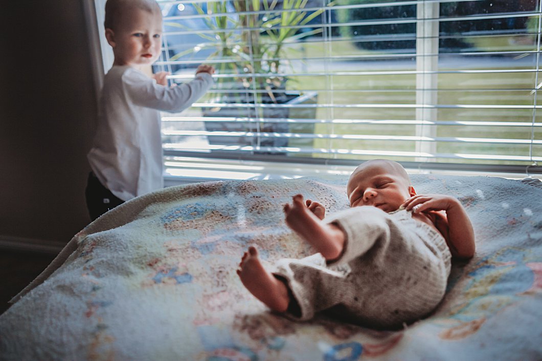 Toddler-looking-out-window-gets-distracted-by-newborn-brother-making-some noises-nearby-gold-coast-in-home-newborn-photography