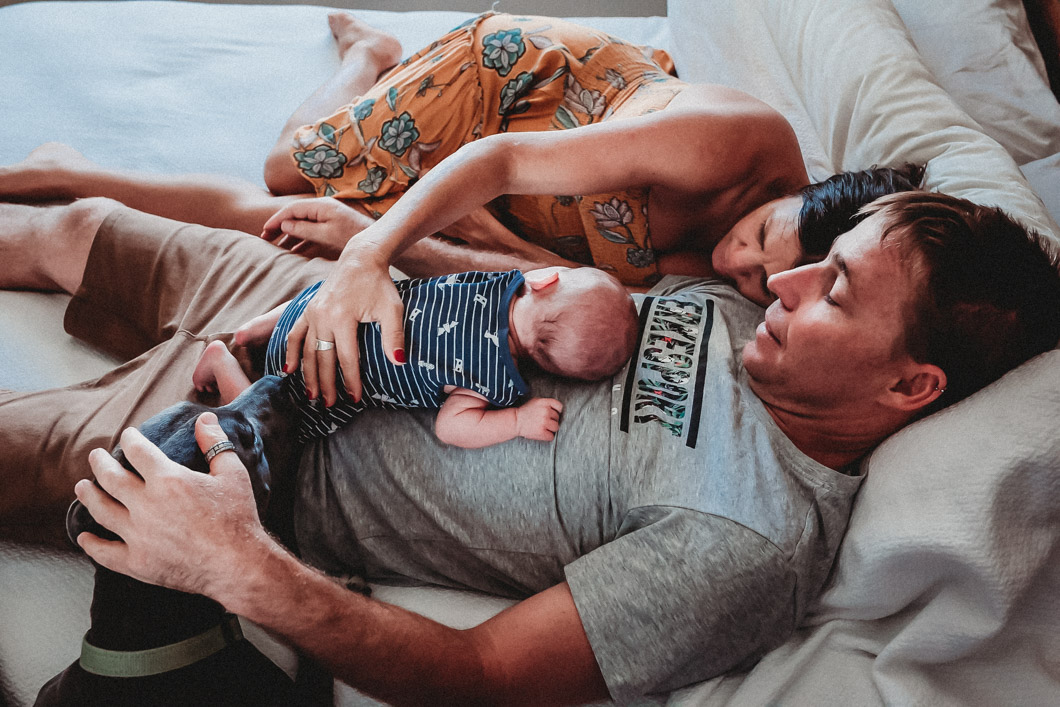 New-parents-resting-on-the-bed-with-their-newborn-son-snuggled-into-dads-chest-whilst-their-pet-dog-comes-close-for-a sniff-of-the-baby