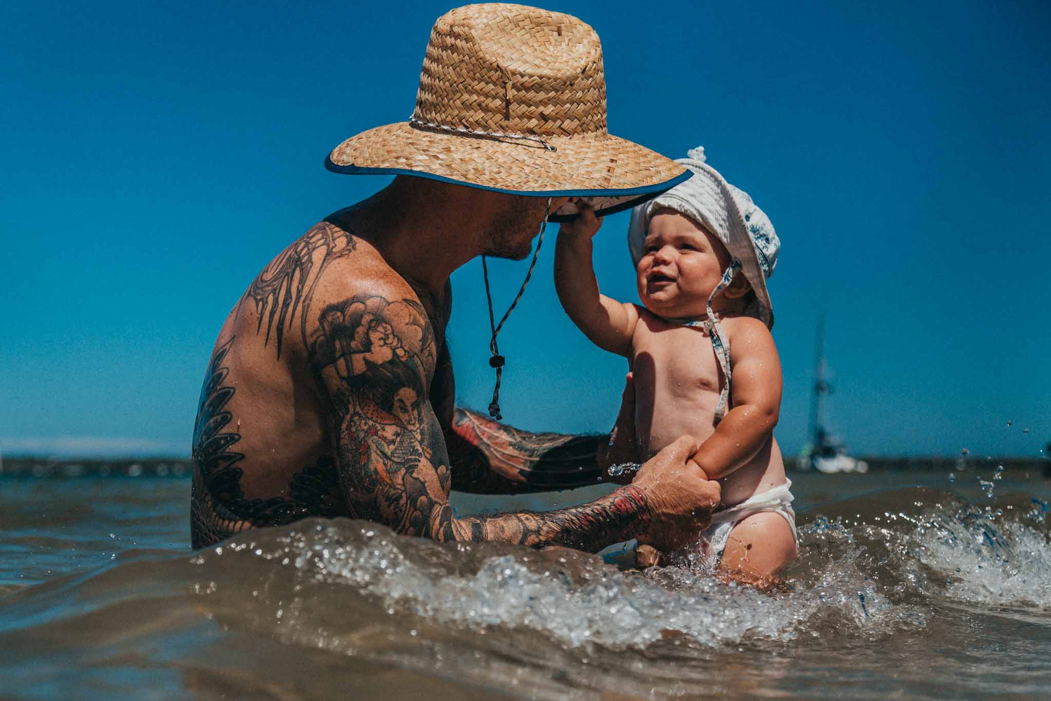 father-playing-with-baby-in-the-beach-with-baby-grabbing-onto-dads-hat-as-a-wave-approaches