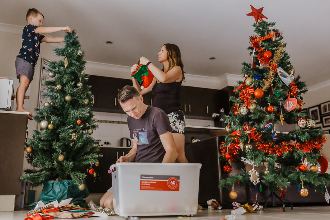 action shot of a family putting up their Christmas trees with a Chile on the counter reaching the higher spots, dad choosing ornaments from a box and mum decorating the other side of the tree