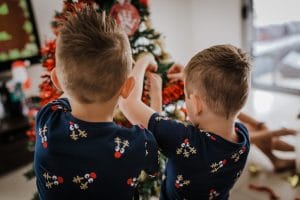 2 young brothers standing side by side as they hang Christmas decorations onto the xmas tree