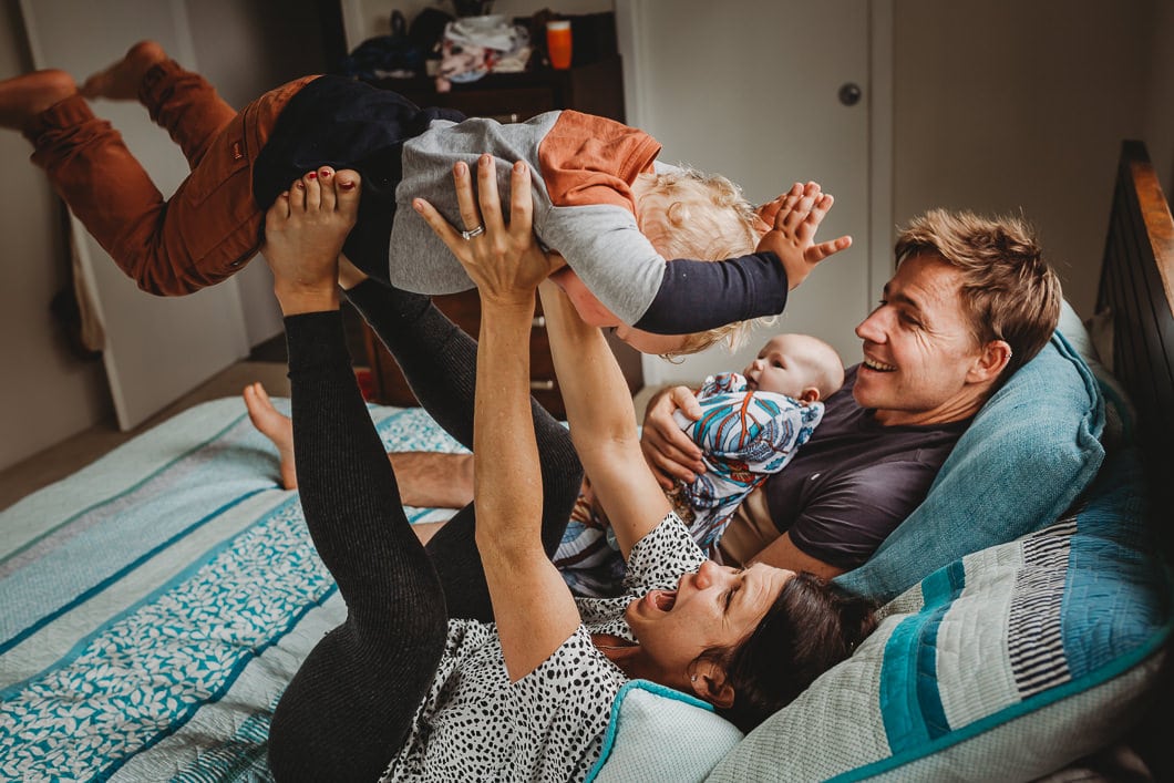 Mum and Dad on the bed playing with toddler son who is flying by putting his belly on mums feet as she lifts him whilst dad is holding on to newborn baby sister