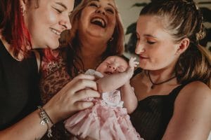 New mum proudly sharing a moment with her sister and mum whilst introducing baby girl. all are laughing with grandma throwing her head back in delight