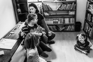 Mother and three daughters in home schooling ring with elder daughters sitting on the bench with mum and toddler on the ground with legs crossed after completing the periodic tables makes an awesome scrunch face at the camera hysterical