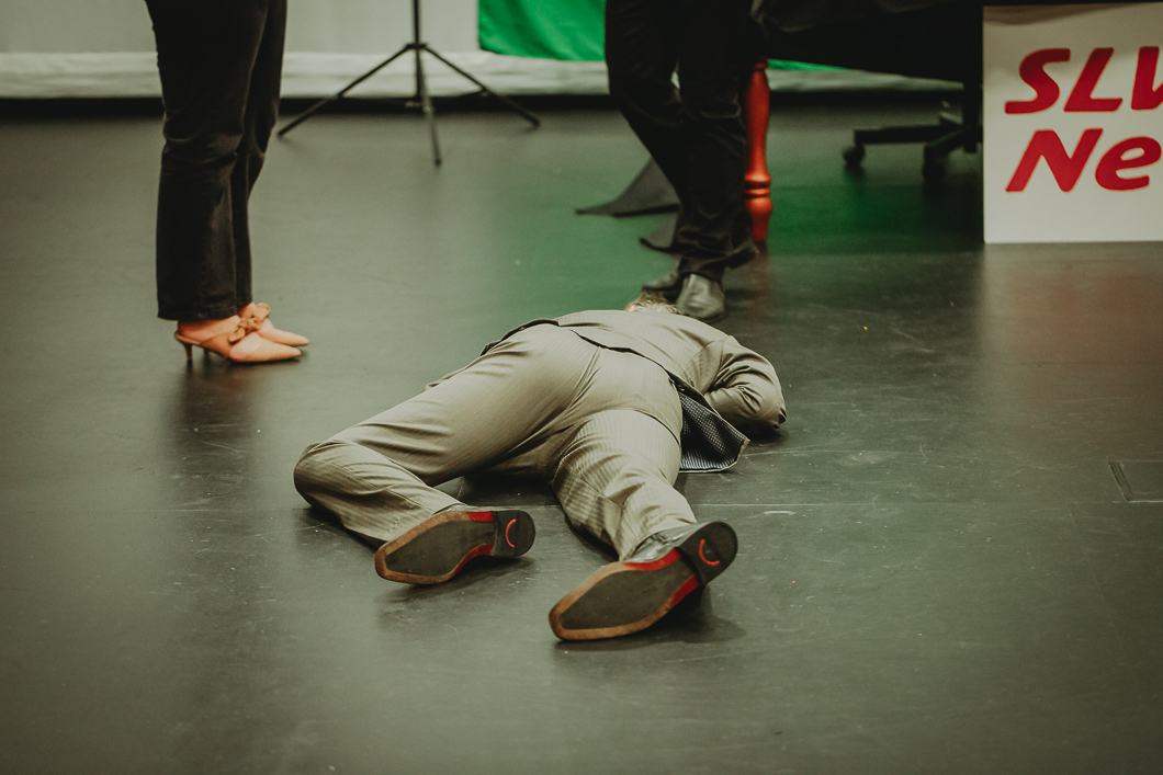 Image of a man in a play passed out on the stage floor viewing from his feet up to his head