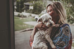 woman-standing-infront-of-large-window-cuddling-white-dog-with-smile-on-her-face