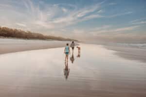 Mother-is-walking-on-the-wet-sand-at-the-Spit-Beach-during-a-sunset-family-beach-photography-session-with-her-large-dog-rodesian-ridgeback-and-child-following-as-the sun-sets-behind-the-beach-lighting-up-the-sky-and-clouds-pink