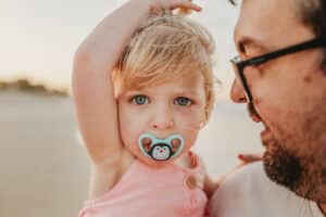 Young-todler-girl-having-family-pictures-at-sunset-on-the-gold-coast-beach-staring-straight-down-camera-lens-with-clear-blue-eyes-as-she-starts to-twist-her-hair-and-suck-on-pacifier-as-she-becomes-tired