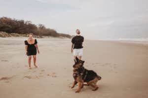 Mum-tossing-the-ball-on-spit-beach-gold-coast-with-adult-son-and-beautful-dog-on-a-windy-day-with-mum-laughing-as-dog-and-son-wait-for-ball-caught-up-in-the-wind